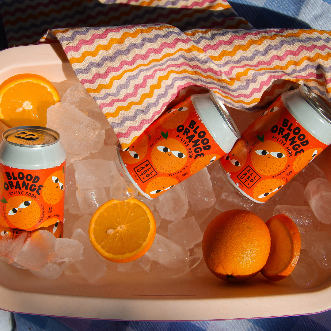 Cans of Blood Orange A-Live Soda on ice with fresh orange slices, styled in a summer picnic setup