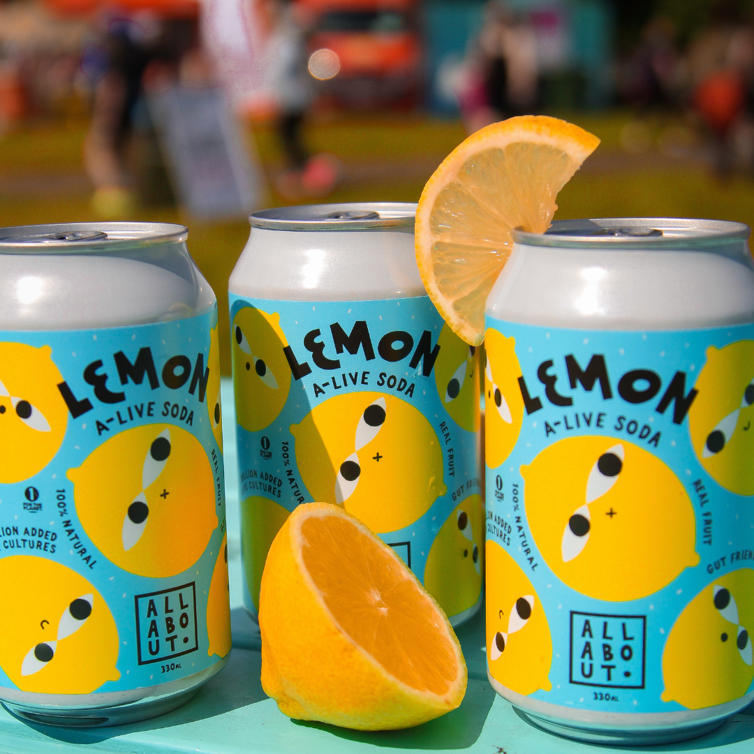 Three cans of All About Kombucha Lemon A-Live Soda with fresh lemon slices, displayed outdoors in natural sunlight