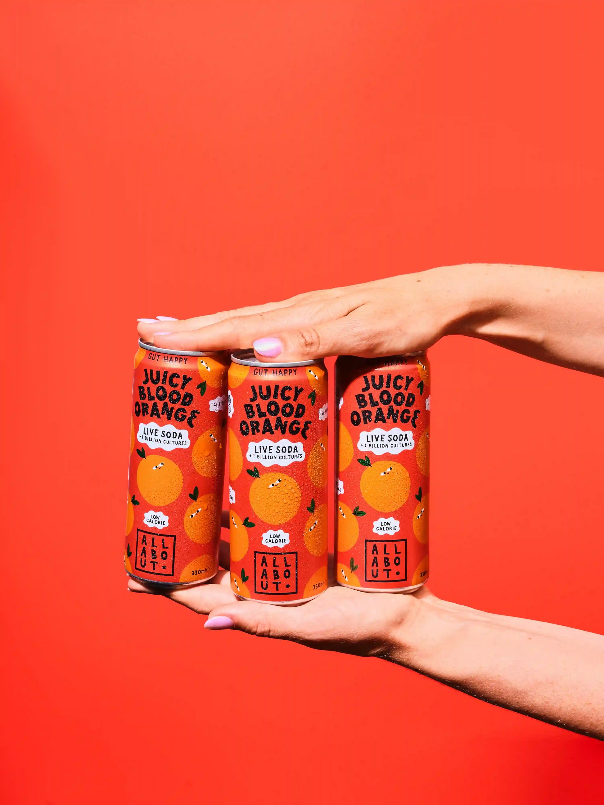 Three cans of Juicy Blood Orange Live Soda held in two hands against a solid bright red background.