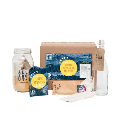 All About Kombucha Deluxe Brewing Kit, including a kombucha brewing guidebook, home brewing bible, glass brewing jar, labeled ingredients pack, reusable cloth, clear swing-top bottle with a tag, and a kraft paper box with detailed product labeling.