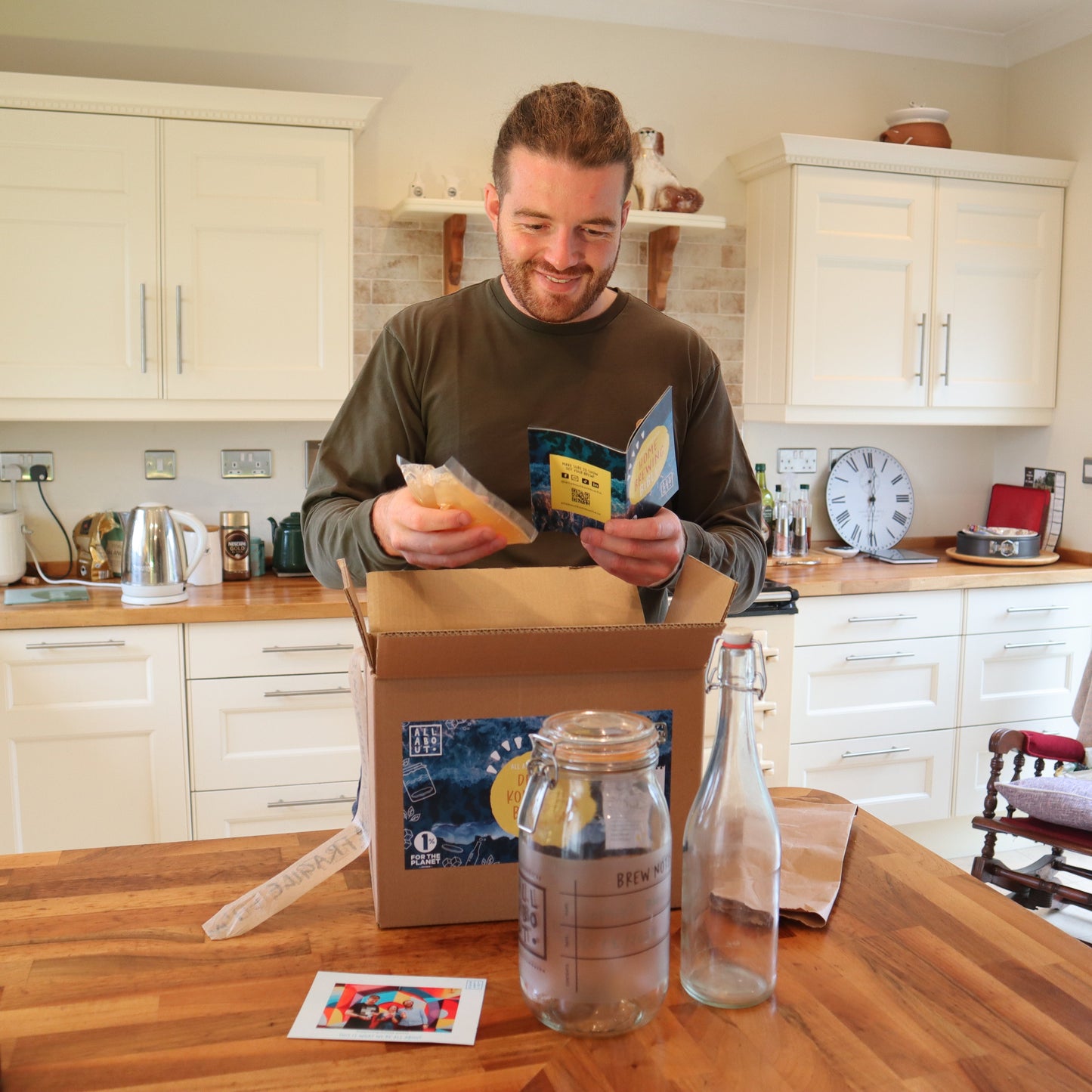 Man smiling while unboxing the All About Kombucha home brewing kit in a bright kitchen, holding ingredients and a brewing guide with a glass jar and bottle on the counter
